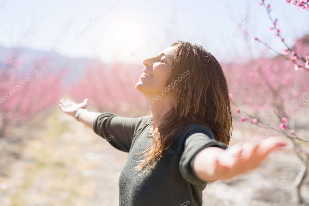 Beautiful middle age woman in the middle of pink peach flowers and trees smiling cheerful with open arms enjoying sunbathe on sunny day