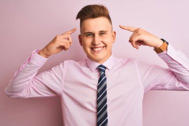 Genç yakışıklı işadamı gömlek ve kravat izole pembe arka plan üzerinde duran iki eliyle parmak, harika bir fikir ya da düşünce, iyi bellek ile baş gösteren giyiyor