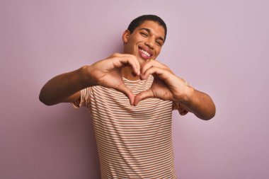 Genç yakışıklı Arap adam çizgili t-shirt izole pembe arka plan üzerinde duran giyen aşk kalp sembolü ve elleriyle şekil gösteren gülümseyerek. Romantik kavram.