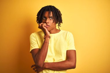 Rahat t-shirt giyen dreadlocks ile Afro adam izole sarı arka plan üzerinde duran stresli ve ağız tırnak ısırma elleri ile sinir görünümlü. Anksiyete sorunu.
