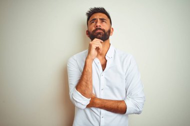 Zarif gömlek giyen genç hintli adam çenesi üzerinde el ile izole beyaz arka plan üzerinde duran soru, dalgın ifade düşünüyor. Düşünceli bir yüzle gülümsüyor. Şüphe kavramı.