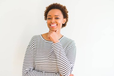 Beyaz arka plan üzerinde çizgili kazak giyen genç güzel Afrikalı Amerikalı kadın çapraz silah ve el çene üzerinde yükseltilmiş ile gülümseme ile kamera emin bakıyor. Pozitif düşünme.