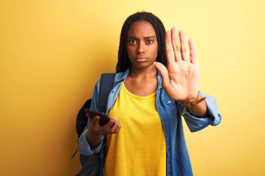 Afro-Amerikan öğrenci kadın akıllı telefon kullanarak izole edilmiş sarı arka planda duruyor. Açık elle dur işareti yapıyor. Ciddi ve kendinden emin bir ifadeyle, savunma hareketi yapıyor.