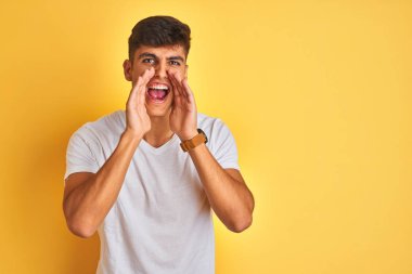 İzole sarı arka plan üzerinde duran beyaz t-shirt giyen genç hintli adam ağız üzerinde elleri ile yüksek sesle kızgın bağırArak