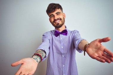 Dövme mor gömlek ve izole beyaz arka plan üzerinde papyon giyen genç adam sarılmak için açık kollarla gülümseyen kameraya bakarak. Mutluluğu kucaklayan neşeli ifade.