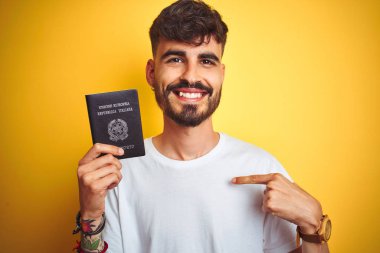 Kendine işaret sürpriz yüzü ile izole sarı arka plan üzerinde İtalya İtalyan pasaportu giyen dövme ile genç adam