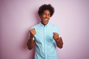 Afro saçlı, mavi gömlek giyen genç amerikalı adam izole pembe arka plan üzerinde duran çok mutlu ve heyecanlı kolları kaldırdı ile kazanan jest yapıyor, gülümseyerek ve başarı için çığlık. Kutlama konsepti.