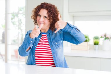 Kıvırcık saçlı orta yaş yaşlı kadın evde kot ceket giyen gülümseyerek telefon jesti yaparak ve sizi işaret ediyor. Beni ara.