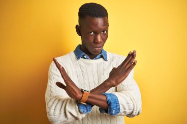 Izole sarı arka plan reddi ifade geçiş kolları üzerinde denim gömlek ve beyaz kazak giyen Afro-amerikan adam olumsuz işaret yapıyor, kızgın yüz
