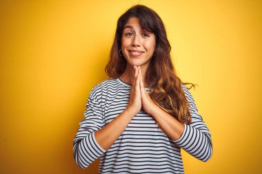 Çizgili t-shirt giyen genç güzel kadın birlikte emin gülümseyerek af isteyen elleri ile dua yelllow izole arka plan üzerinde duran.