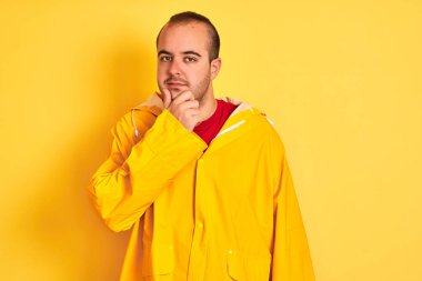 Izole sarı arka plan üzerinde duran yağmurluk giyen genç adam çapraz kolları ve çene üzerinde kaldırdı el ile gülümseme ile kameraya emin bakıyor. Olumlu düşünme.