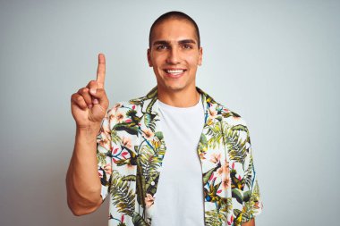Genç yakışıklı adam tatillerde beyaz arka plan üzerinde Hawaii gömlek giyen gösteren ve kendinden emin ve mutlu gülümserken parmak sayısı bir ile işaret.