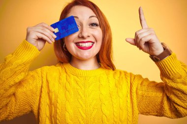 Genç güzel redhead kadın sarı izole arka plan üzerinde kredi kartı tutan mutlu yüz, bir numaralı ile bir fikir veya soru işaret parmağı ile şaşırttı