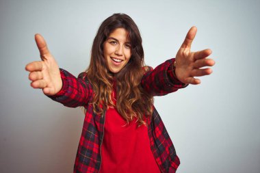 Kırmızı tişört ve ceket giyen genç güzel kadın beyaz izole arka plan üzerinde duran sarılmak için açık kollarla gülümseyen kameraya bakıyor. Mutluluğu kucaklayan neşeli ifade.