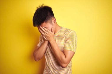 Genç Asyalı Çinli adam feryat ederken elleri ile yüz kapsayan üzgün ifade ile izole sarı arka plan üzerinde duran t-shirt giyiyor. Depresyon kavramı.