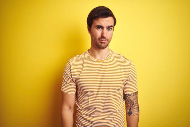 Izole sarı arka plan şüpheci ve sinir üzerinde çizgili t-shirt giyen dövme ile genç yakışıklı adam, sorun nedeniyle üzgün kaşlarını çatma. Negatif kişi.