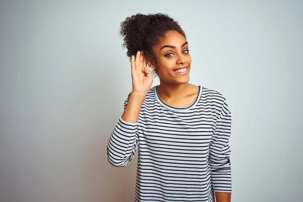 African american woman wearing navy striped t-shirt standing over isolated white background smiling with hand over ear listening an hearing to rumor or gossip. Deafness concept.