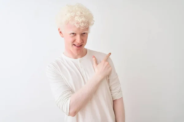 Young albino blond man wearing casual t-shirt standing over isolated white background cheerful with a smile of face pointing with hand and finger up to the side with happy and natural expression on face