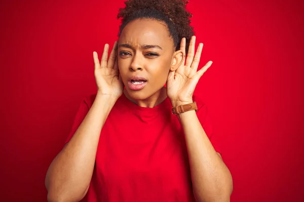 Young beautiful african american woman with afro hair over isolated red background Trying to hear both hands on ear gesture, curious for gossip. Hearing problem, deaf