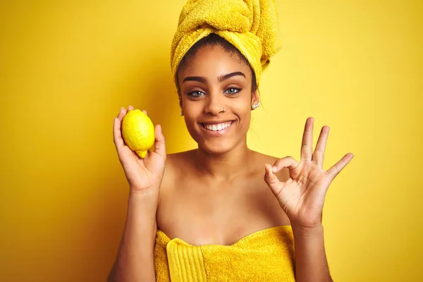 Afro american woman wearing towel after shower holding lemon over isolated yellow background doing ok sign with fingers, excellent symbol