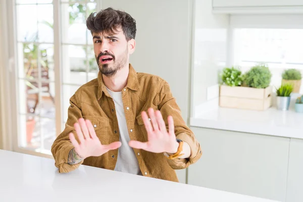 Young man wearing casual jacket sitting on white table afraid and terrified with fear expression stop gesture with hands, shouting in shock. Panic concept.