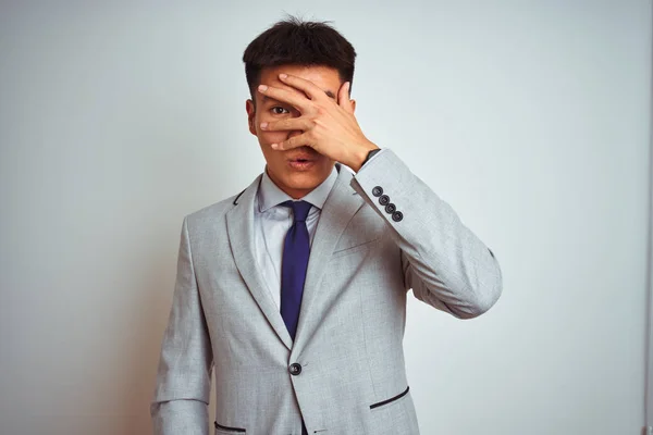 Asian chinese businessman wearing suit and tie standing over isolated yellow background peeking in shock covering face and eyes with hand, looking through fingers with embarrassed expression.