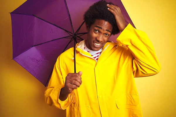 Afro american man wearing rain coat and umbrella standing over isolated yellow background stressed with hand on head, shocked with shame and surprise face, angry and frustrated. Fear and upset for mistake.