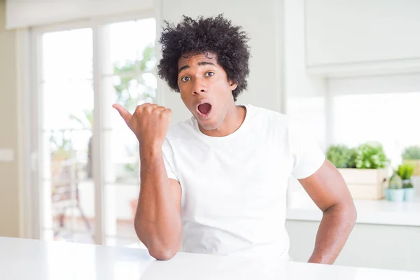 カジュアルな白いTシャツを着た若いアフリカ系アメリカ人男性が自宅に座って驚き 手指で横に指差し 口を開いた表情 — ストック写真