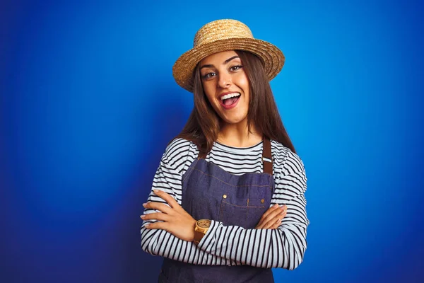 Young beautiful baker woman wearing apron and hat standing over isolated blue background happy face smiling with crossed arms looking at the camera. Positive person.