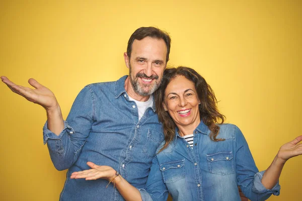 Güzel orta yaş çift birlikte izole sarı arka plan üzerinde duran gülümseyerek her iki elaçık avuç içi gösteren, sunan ve reklam karşılaştırma ve denge