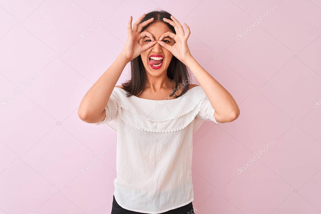 Beautiful chinese woman wearing white t-shirt standing over isolated pink background doing ok gesture like binoculars sticking tongue out, eyes looking through fingers. Crazy expression.