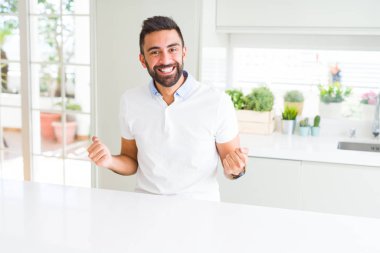 Evde yakışıklı İspanyol adam Casual beyaz t-shirt sürpriz kutluyor ve silah yükseltilmiş ve açık gözleri ile başarı için hayran. Kazanan kavramı.
