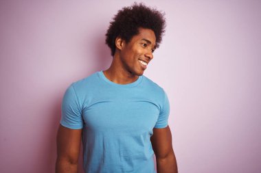 Afro saçlı afro saçlı, izole pembe arka plan üzerinde duran mavi t-shirt giyen Afrikalı Amerikalı adam yüzünde gülümseme, doğal ifade ile yan bakıyor. Gülmekten emin.