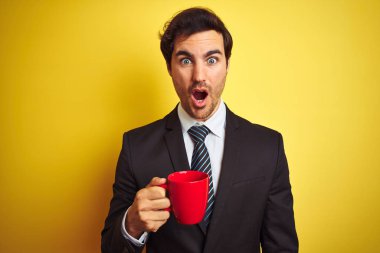 Izole sarı arka plan üzerinde kırmızı fincan kahve içen genç yakışıklı işadamı sürpriz bir yüz ile şok korkmuş, korku ifade korku ve heyecanlı