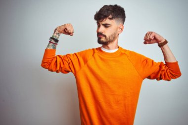 Dövmeli genç adam turuncu kazak giyen izole beyaz arka plan üzerinde duran kol kasları gururla gülümseyen gösteren. Fitness konsepti.