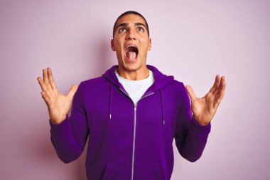 Genç yakışıklı adam pembe izole arka plan deli ve deli bağırarak üzerinde mor sweatshirt giyen ve agresif ifade ve kollar kaldırdı ile bağırıyor. Hayal kırıklığı kavramı.