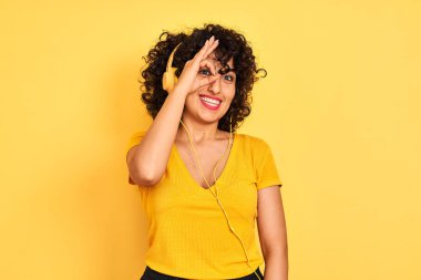 Kıvırcık saçlı Arap kadın izole sarı arka plan üzerinde kulaklık kullanarak müzik dinlerken el gülümseyerek ok jest yapıyor, mutlu yüz ile parmaklardan bakan göz.