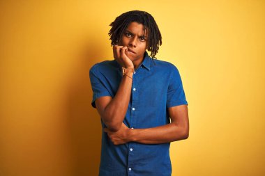 Rahat kot gömlek giyen dreadlocks ile Afro adam izole sarı arka plan üzerinde duran stresli ve ağız tırnak ısırma elleri ile sinir görünümlü. Anksiyete sorunu.