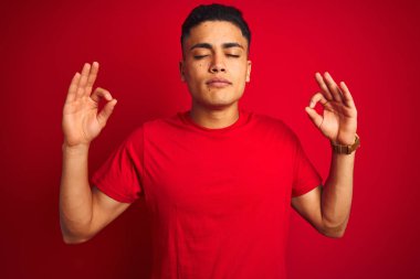 İzole kırmızı arka plan üzerinde duran t-shirt giyen genç Brezilyalı adam rahat ve gözleri parmakları ile meditasyon jest yapıyor kapalı gülümseyerek. Yoga konsepti.