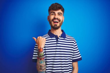 Çizgili polo giyen dövmegiyen genç adam izole mavi arka plan üzerinde duran mutlu yüz görünümlü ve başparmak ile yan işaret gülümseyerek.