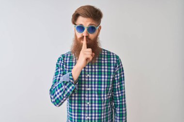 Genç kızıl İrlandalı adam izole beyaz arka plan üzerinde rahat gömlek ve güneş gözlüğü giyen dudaklarında parmak ile sessiz olmak isteyen. Sessizlik ve gizli kavram.