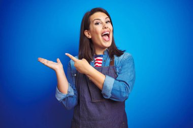 Mavi izole arka plan üzerinde mağaza üniforma önlük giyen genç güzel iş kadını şaşırttı ve el ile sunarken kameraya gülümseyerek ve parmak ile işaret.