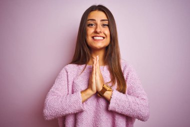 Yalnız pembe arka plan üzerinde duran rahat kazak giyen genç güzel kadın elleri birlikte emin gülümseyerek af isteyen dua.