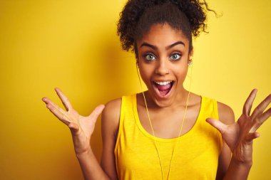 Afro-Amerikan kadın izole sarı arka plan üzerinde kulaklık kullanarak müzik dinlerken deli kutluyor ve kollarını kaldırdı ve açık gözleri heyecan çığlık ile başarı için hayran. Kazanan kavramı