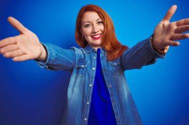 Mavi izole arka plan üzerinde duran denim gömlek giyen genç güzel kızıl saçlı kadın sarılmak için açık kollarla gülümseyerek kameraya bakarak. Mutluluğu kucaklayan neşeli ifade.