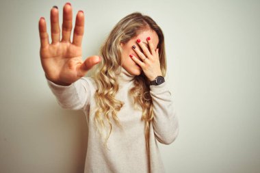 İzole beyaz arka plan üzerinde kış boğazlı kazak giyen güzel kadın elleri ile gözleri kaplayan ve üzgün ve korku ifadesi ile stop jest yapıyor. Utanmış ve olumsuz kavram.