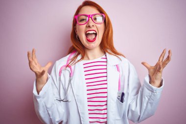 Genç kızıl saçlı doktor kadın izole pembe arka plan üzerinde duran stetoskop kullanarak deli ve deli bağırarak ve agresif ifade ve kollar kaldırdı ile bağırıyor. Hayal kırıklığı kavramı.