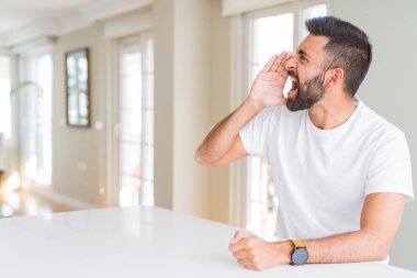 Yakışıklı İspanyol adam evde rahat beyaz t-shirt bağırarak ve ağızda el ile yan yüksek sesle bağırıyor. İletişim kavramı.
