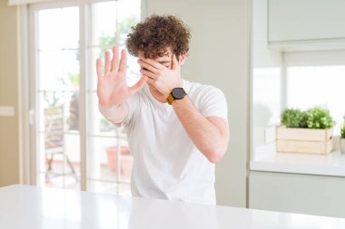 Evde beyaz tişört giyen genç yakışıklı adam elleri ile gözleri kapıyor ve hüzünlü ve korku ifade ile dur jestyapıyor. Utanmış ve olumsuz kavram.