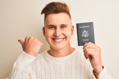 İzole beyaz arka plan üzerinde ABD pasaportu tutan genç yakışıklı adam işaret ve mutlu yüzü gülümseyerek yan başparmak ile gösteren